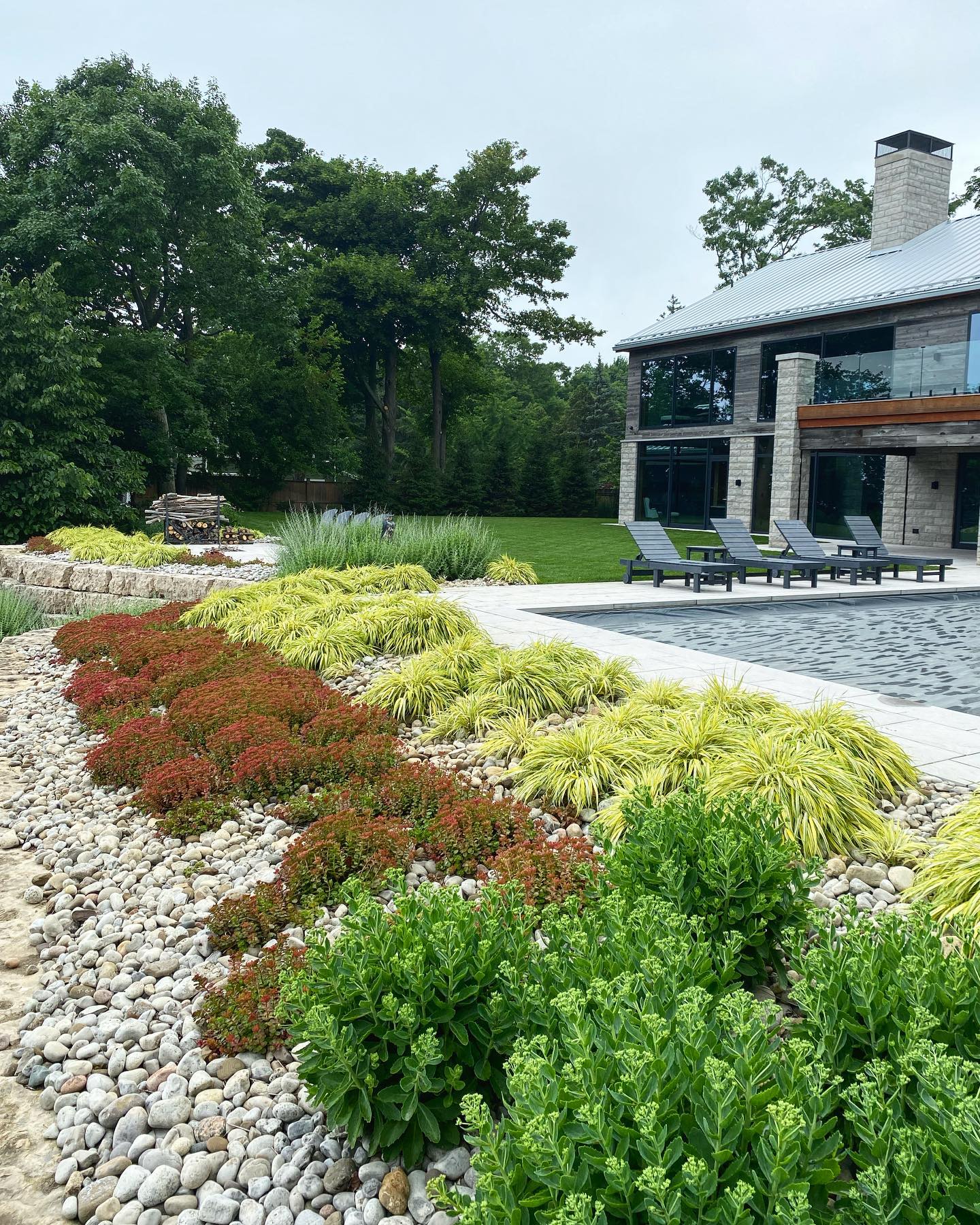 Modern house with large windows and patio, surrounded by landscaped garden featuring various plants and pebble stones. Lush trees in the background.