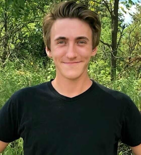 A person wearing a black shirt stands outdoors, surrounded by green foliage and trees, smiling at the camera on a sunny day.