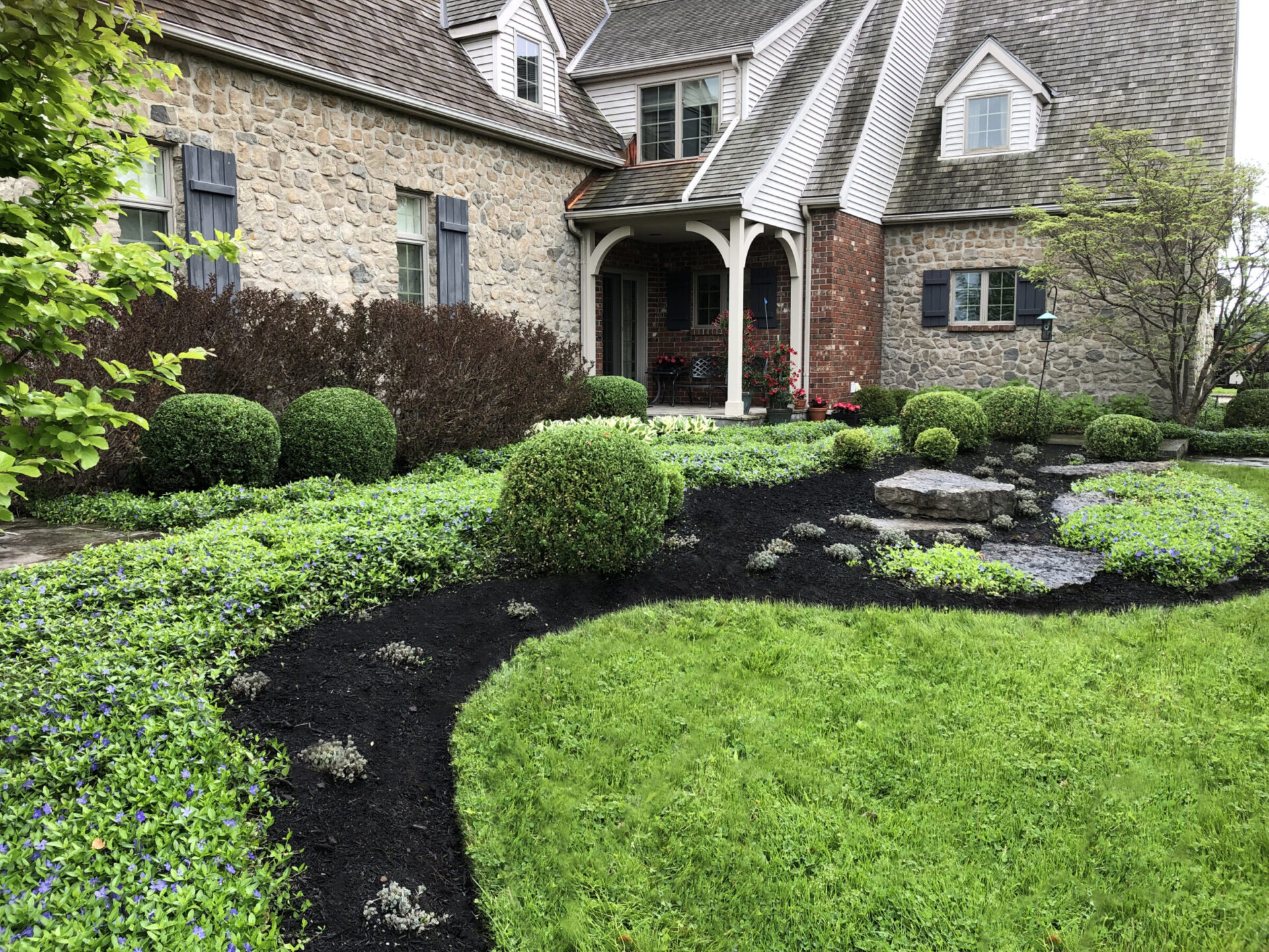 A charming stone house with neatly landscaped gardens, featuring trimmed bushes, vibrant greenery, and a black mulch path leading to the entrance.