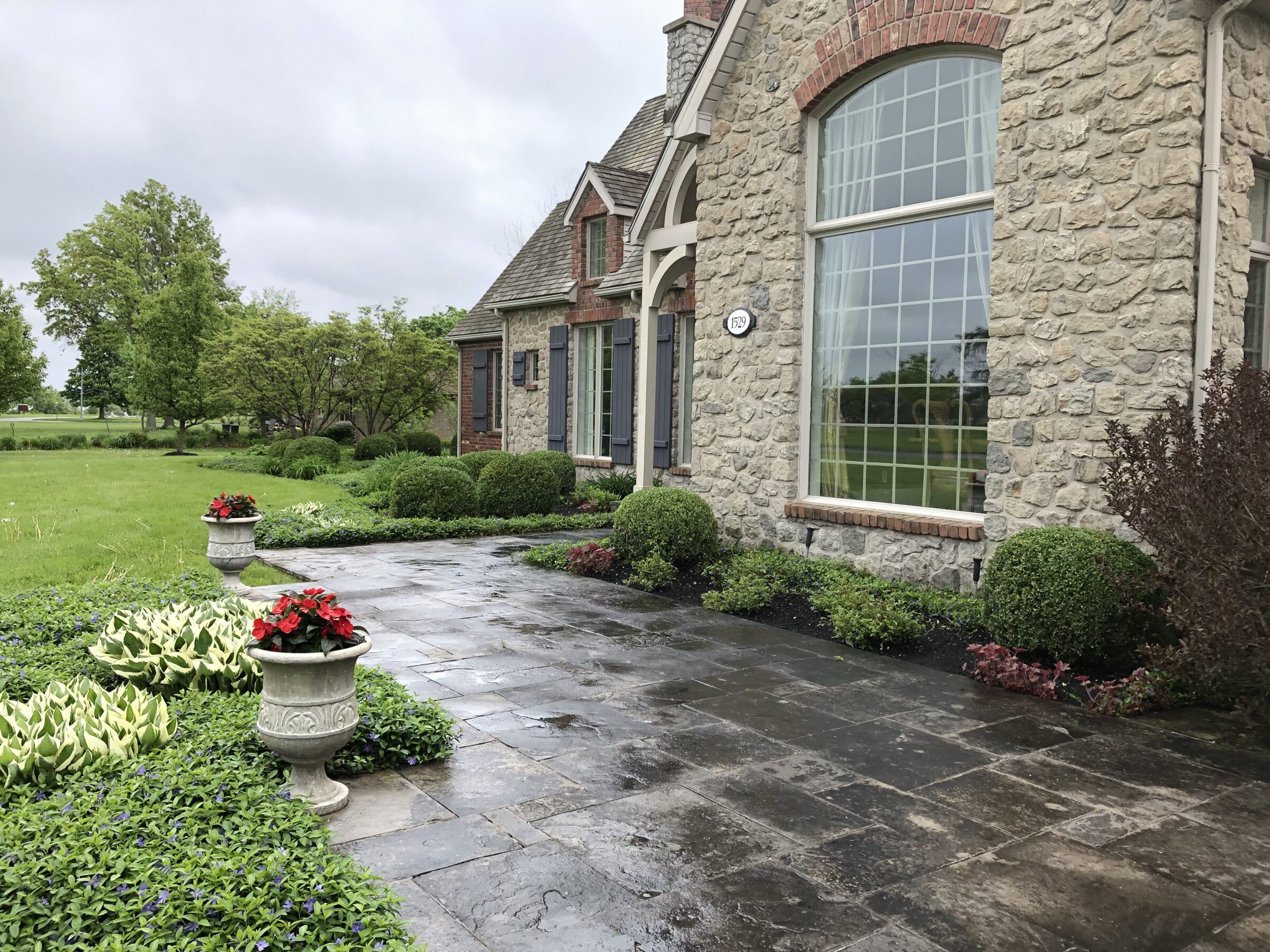 Stone house with large windows, surrounded by lush greenery and manicured shrubs. Decorative planters with red flowers on patio. Overcast sky above.