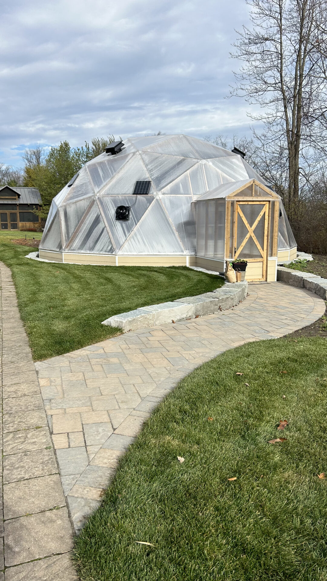 A geodesic dome greenhouse sits on a manicured lawn, surrounded by a stone path, trees, and a smaller wooden structure.