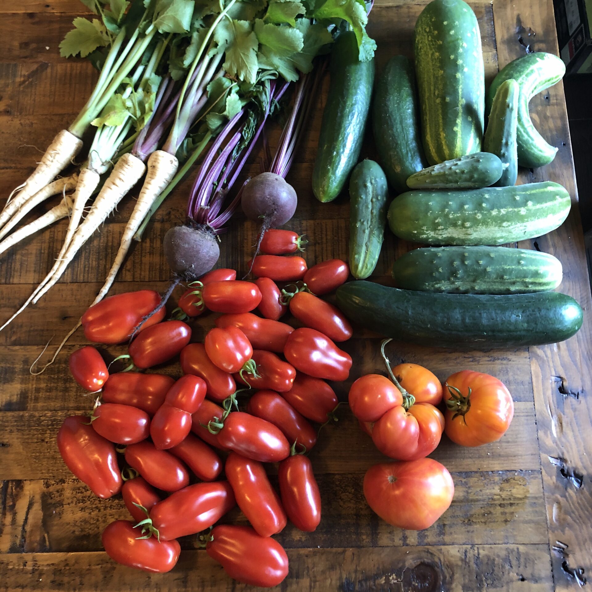 A variety of fresh vegetables, including parsnips, beets, cucumbers, and tomatoes, are displayed on a rustic wooden table.