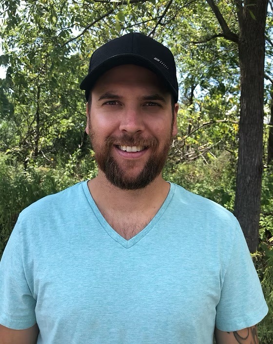 A person in a black cap and teal shirt stands outdoors, smiling. Trees with green leaves fill the background on a sunny day.