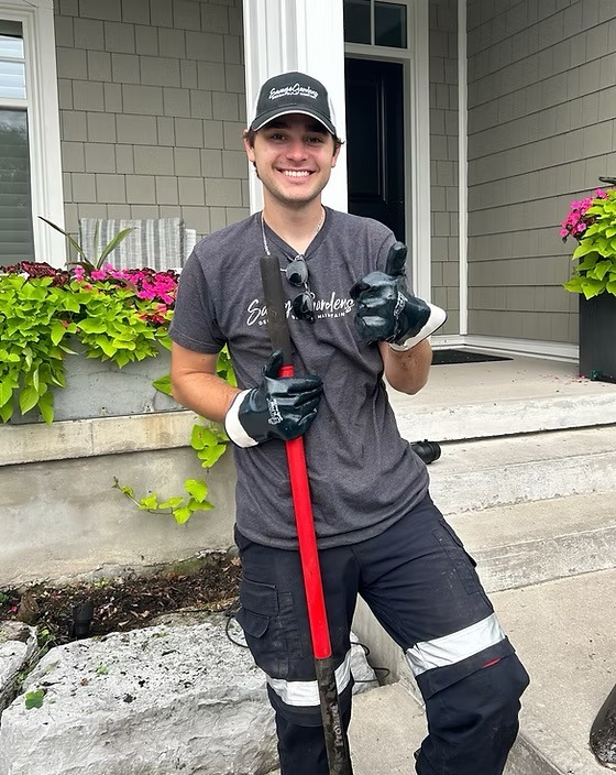 A person smiling, holding a red rake, stands outside a house with potted plants and a gray facade, wearing a cap and gloves.