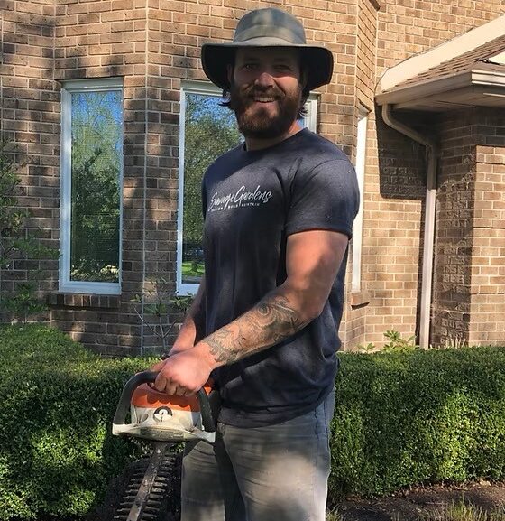 A person in a hat holds a hedge trimmer outside a brick building, standing in a garden with trimmed hedges.