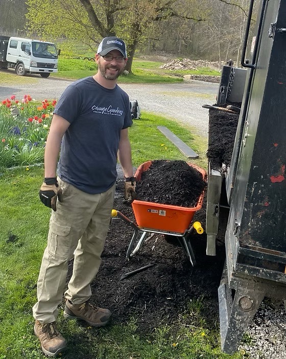 A person stands with a wheelbarrow full of soil near a truck, surrounded by grass and colorful flowers on a sunny day.