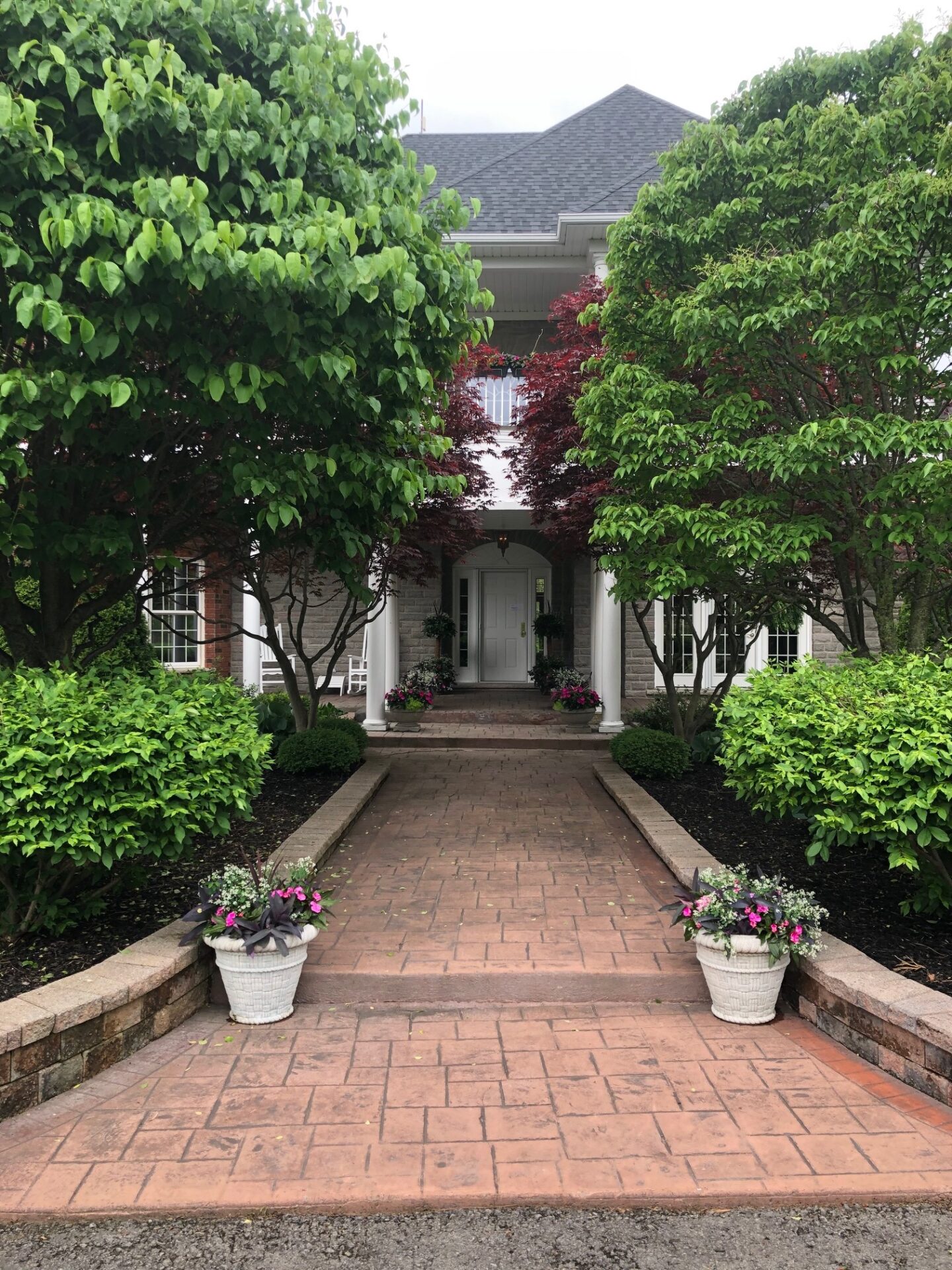 A brick path leads to a house entrance, flanked by lush green trees and potted flowers, creating a peaceful, inviting atmosphere.