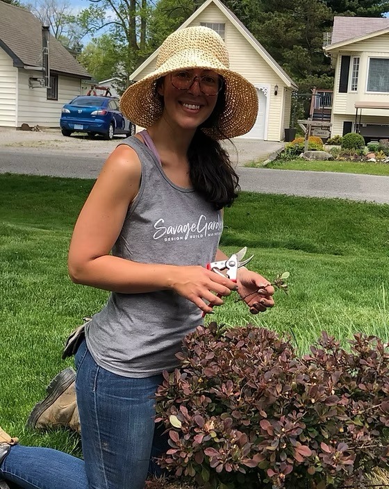 A person wearing a sun hat tends to bushes in a residential garden, smiling. Houses and a blue car are visible in the background.