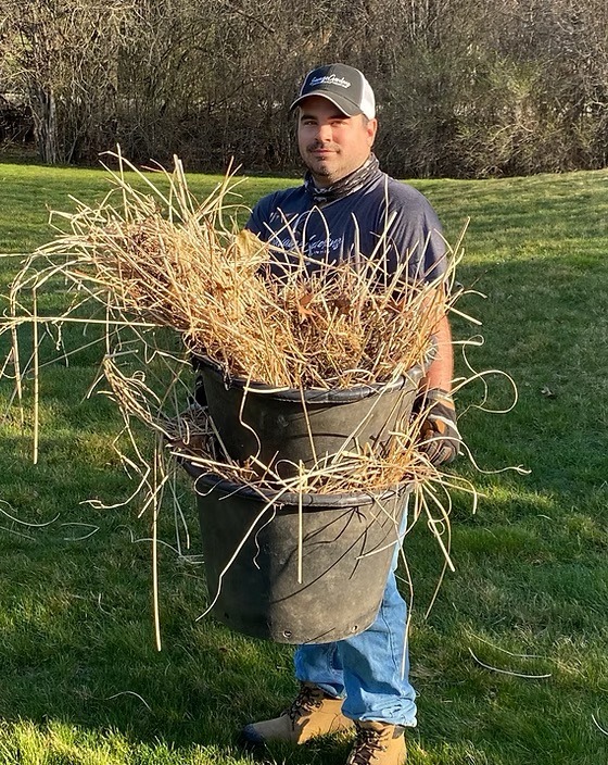 A person in a cap holds a large bucket filled with dry grass on a sunny day, standing on a grassy field.