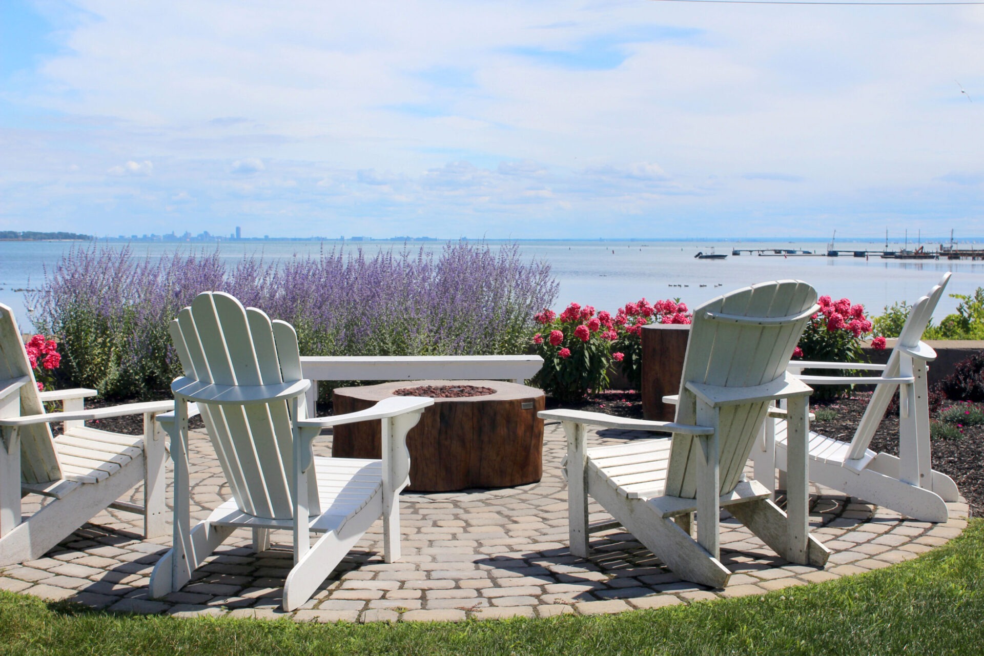 Four Adirondack chairs surround a central fire pit, overlooking a scenic lake with lavender and pink flowers. Skyline visible in distance.