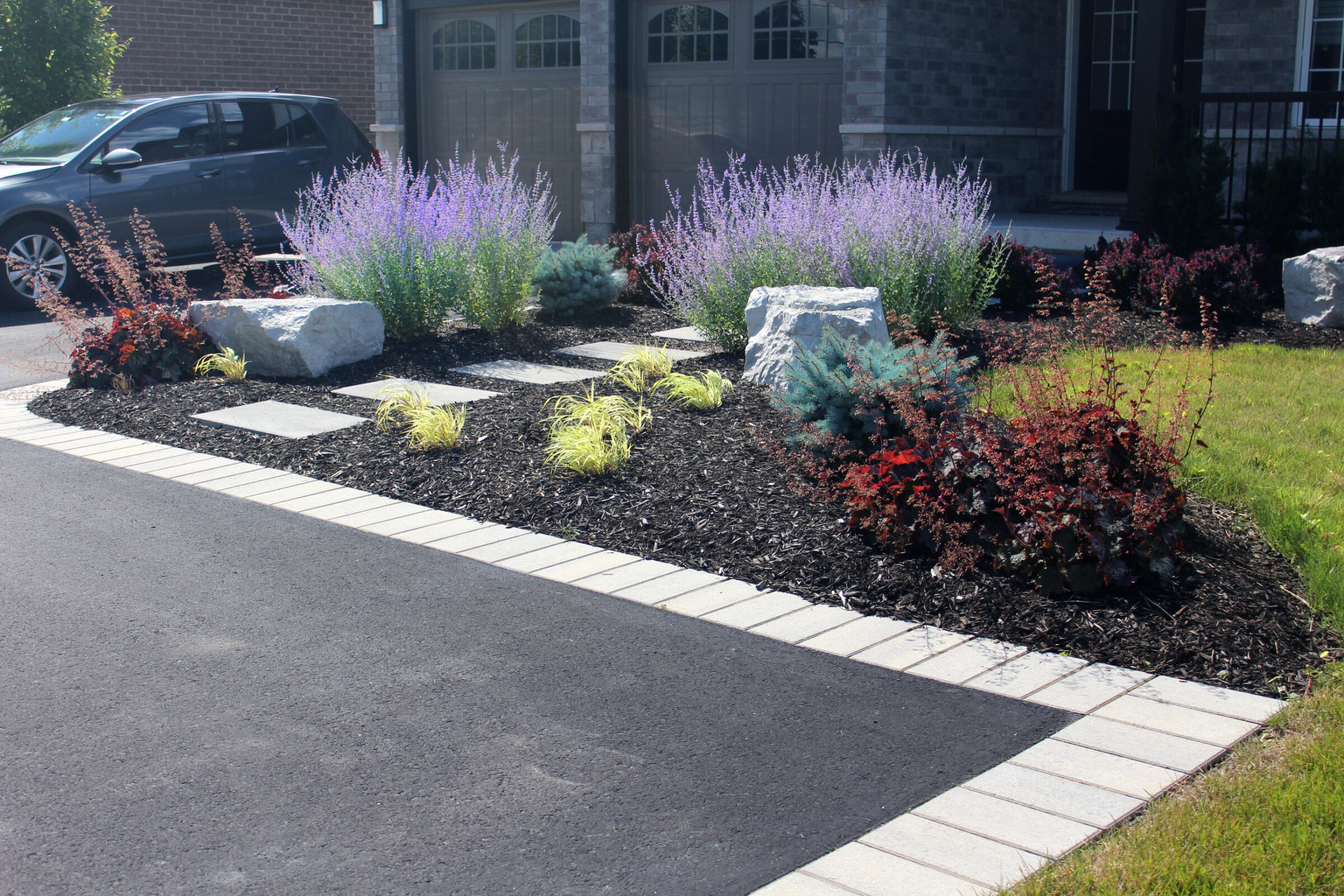 A well-maintained garden with lavender, shrubs, and decorative rocks lines a residential driveway beside a parked car and brick house.