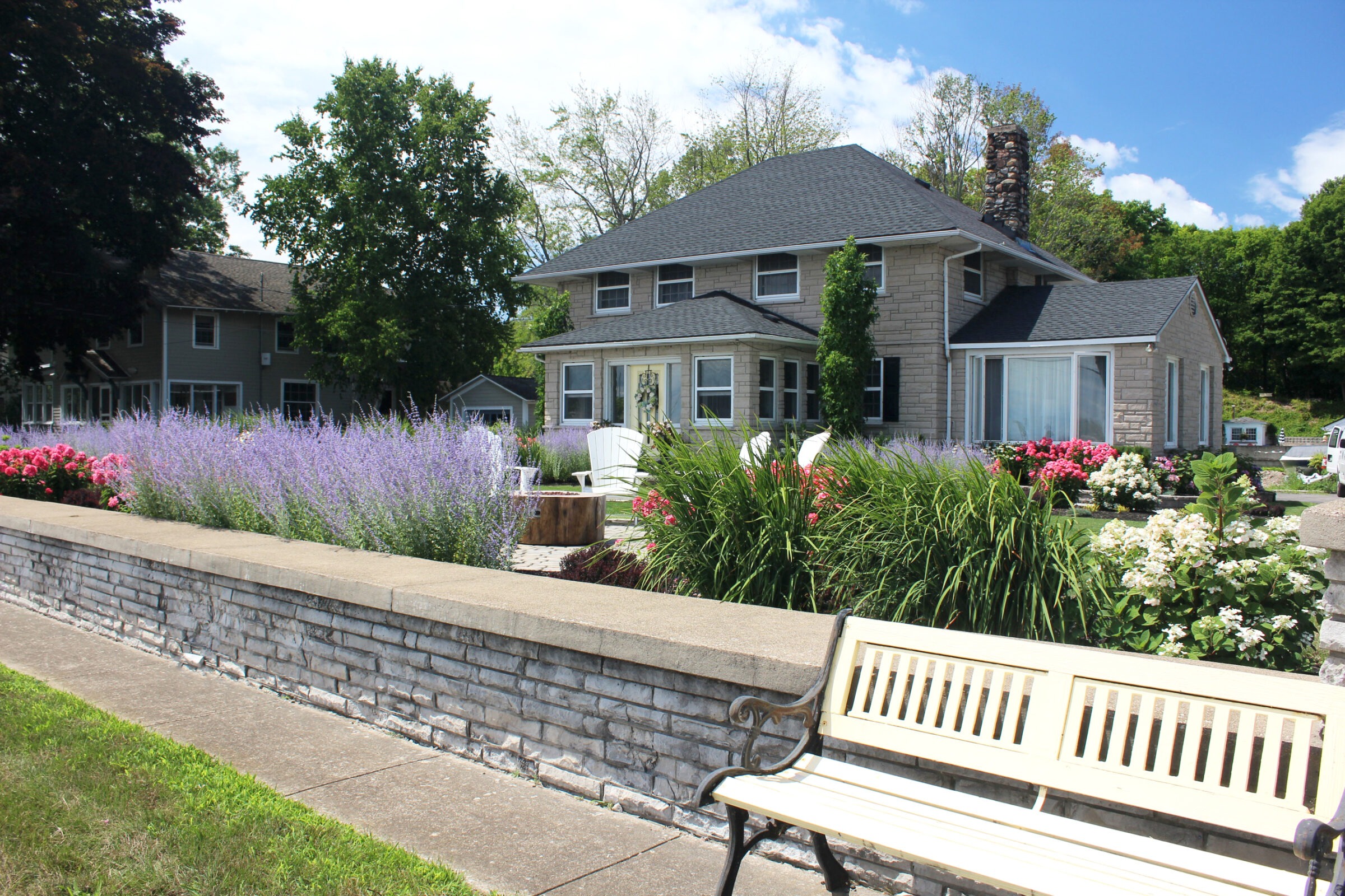 A charming house with a dark roof is surrounded by lush gardens, flowering plants, and a wooden bench along a stone wall.