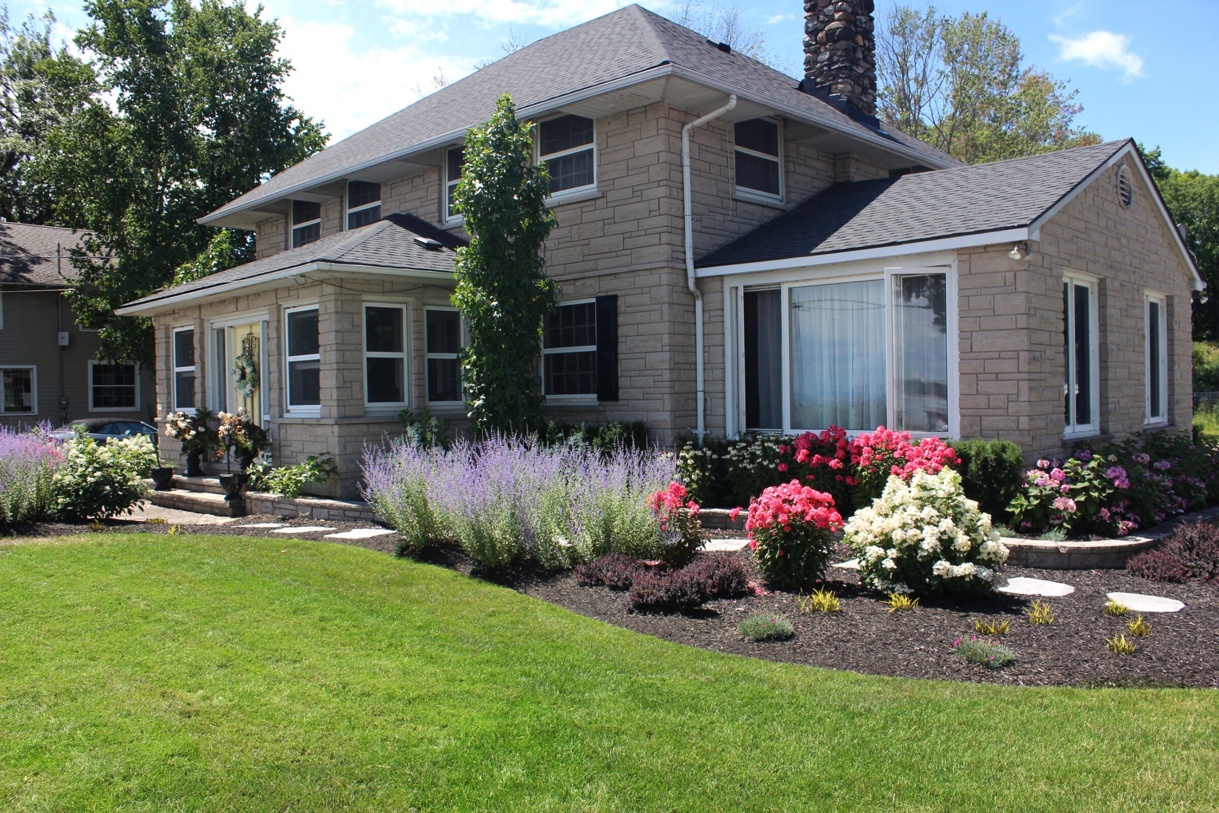 A charming stone house with lush gardens, featuring vibrant flowers and manicured lawns under a clear sky, surrounded by trees and shrubs.