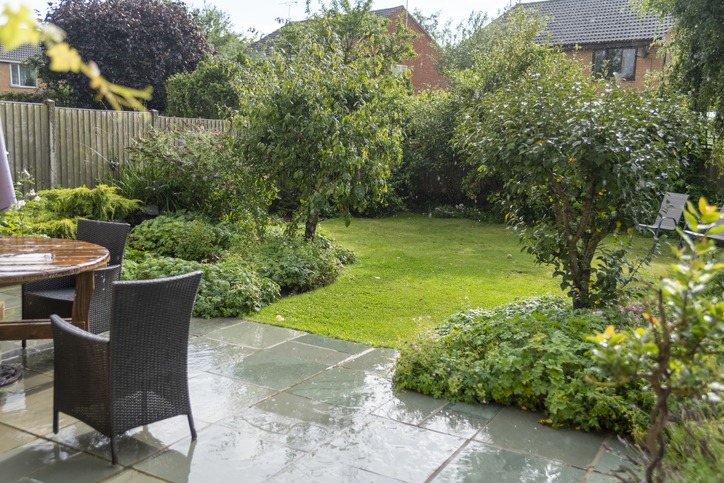 A wet garden with lush greenery, trees, and a patio featuring a wooden table and chairs. Residential houses are visible in the background.