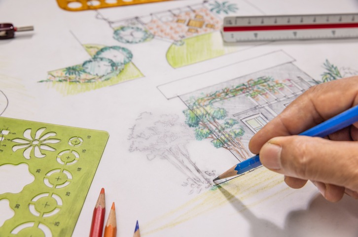 A person sketches architectural designs using colored pencils and stencils. A ruler and additional drawing tools are visible on the table.