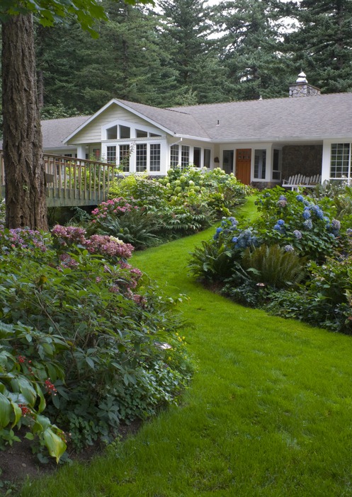 Charming house surrounded by lush garden, vibrant flowers, and dense trees, connected by a wooden bridge. Peaceful, inviting setting with natural beauty.