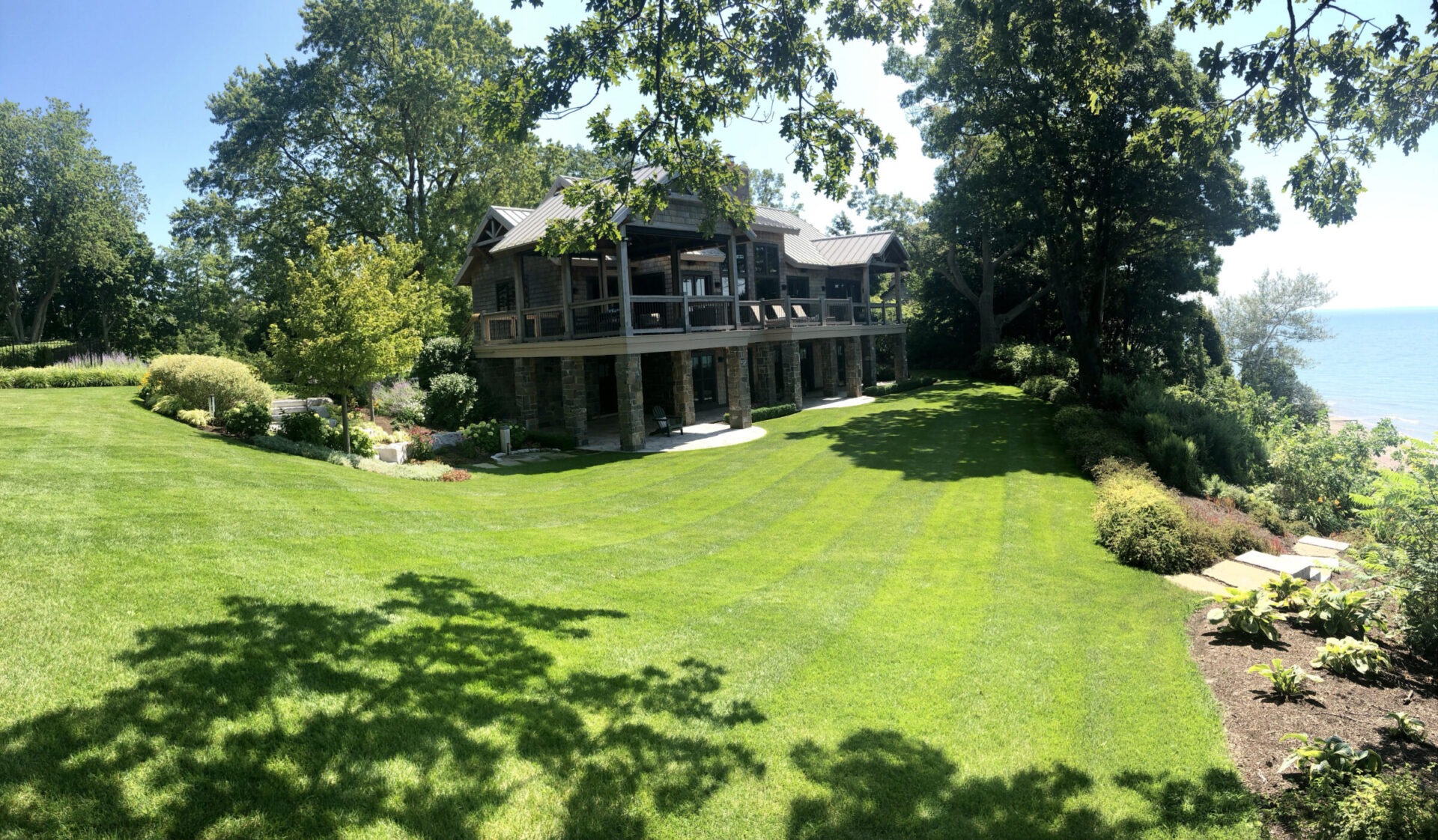 A large house with stone columns sits on a lush green lawn, surrounded by trees, overlooking a body of water under a clear sky.